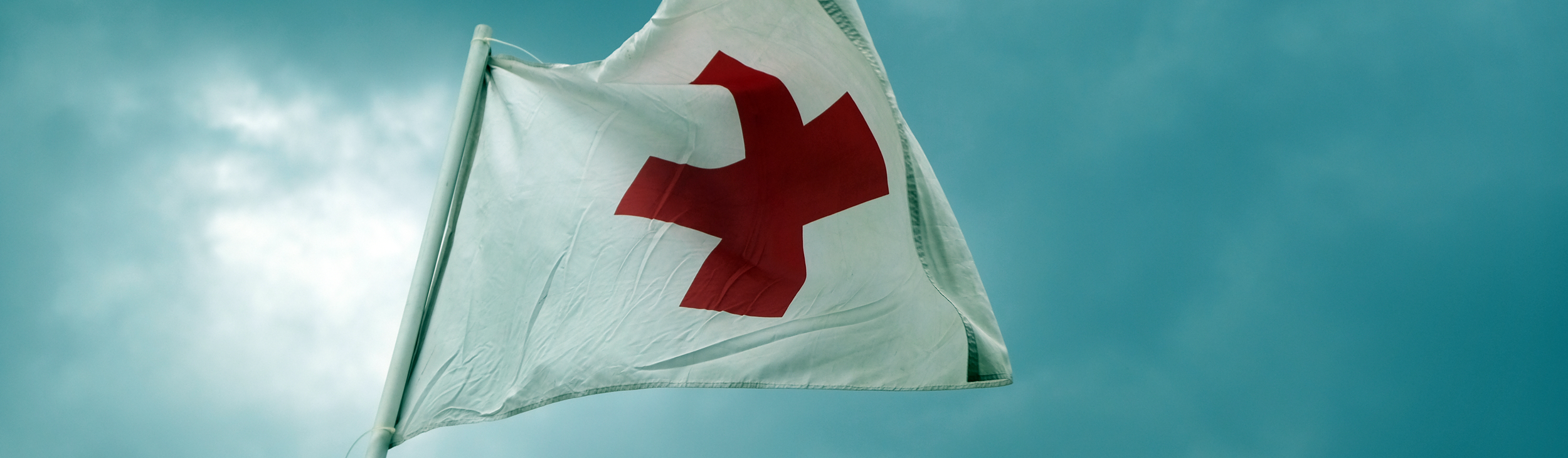 Image of white first aid flag with Red Cross logo, against a cloudy blue sky